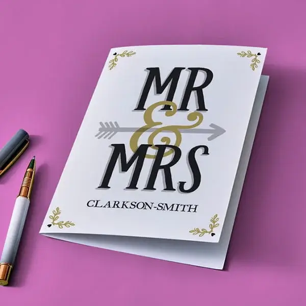 A personalised wedding card with bold Mr & Mrs lettering, an arrow design through the ampersand, and a surname printed below, displayed on a pink background beside two pens.